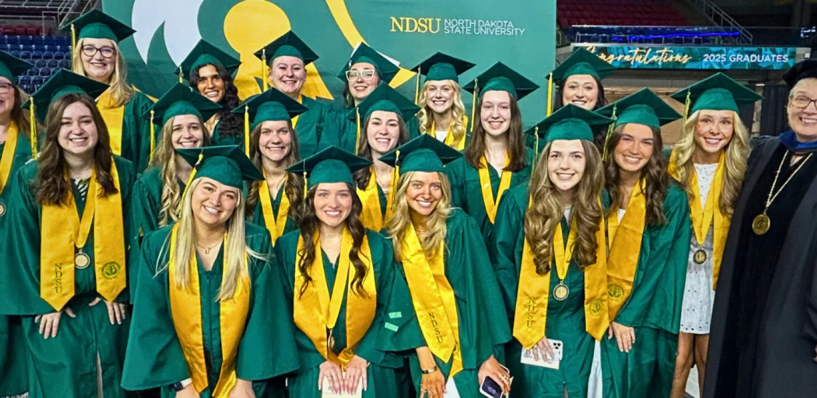 A group of students wearing green graduation gowns with a yellow sash and a faculty member wearing her black gown and black hat.