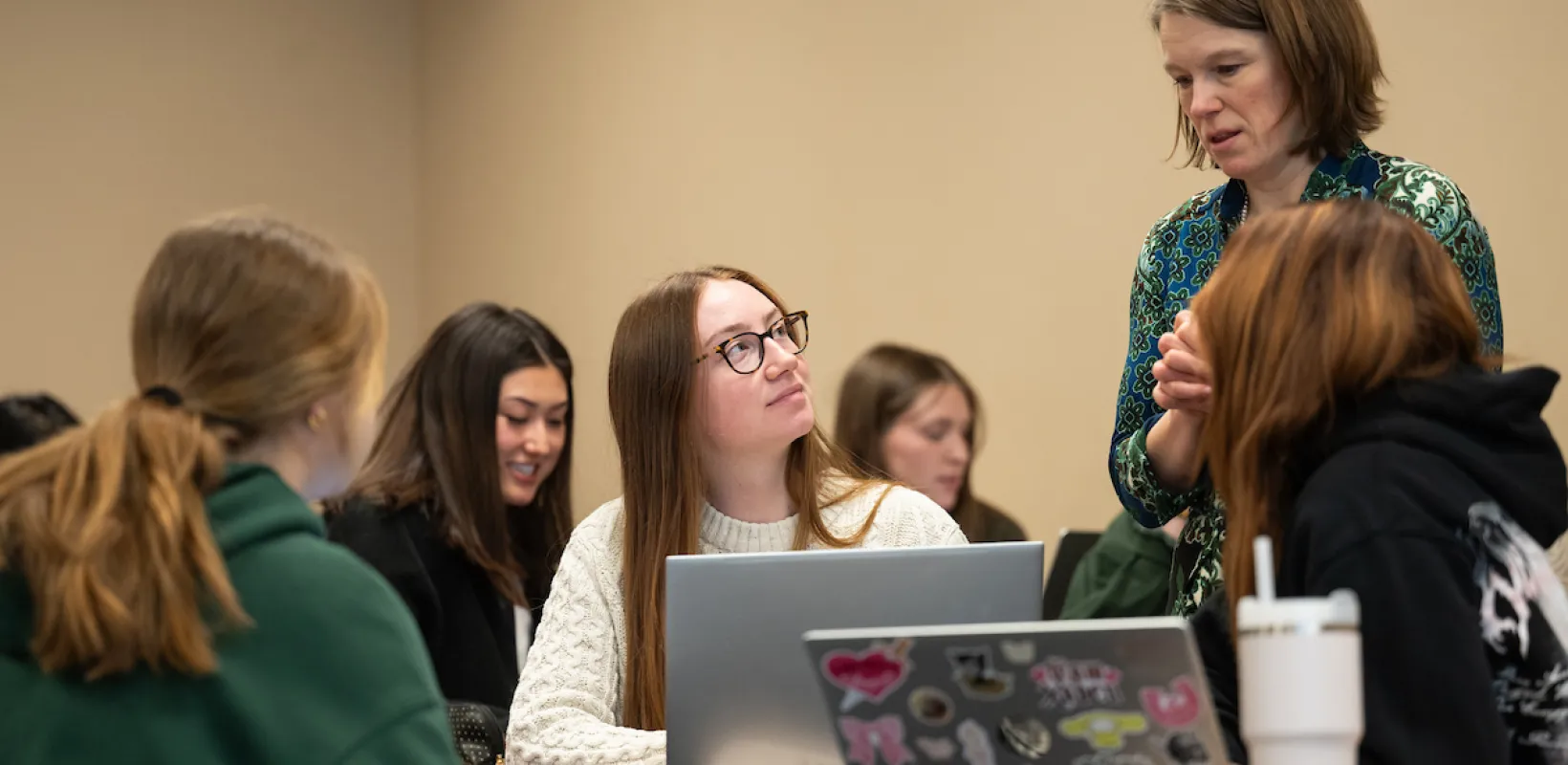 Students visiting with faculty while sitting in class with laptops on desktops.
