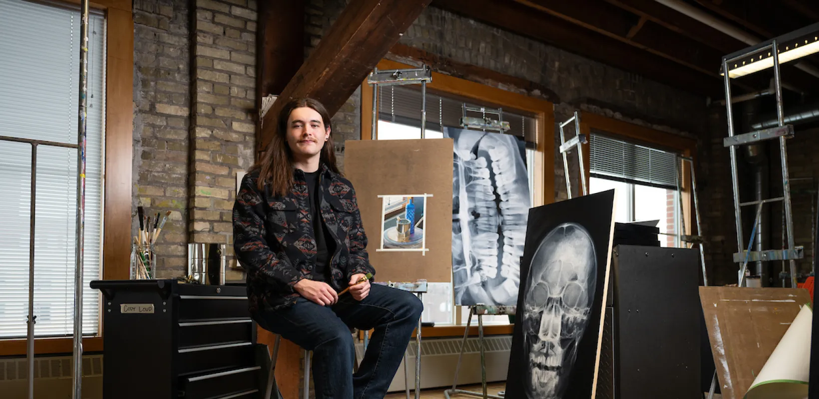 Cody sitting in art studio next to artwork while smiling confidently at camera.