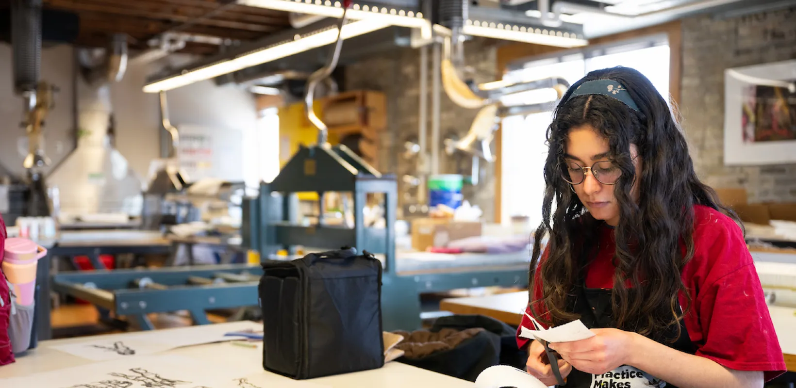 Student cutting paper whiile sitting at table in art studio.