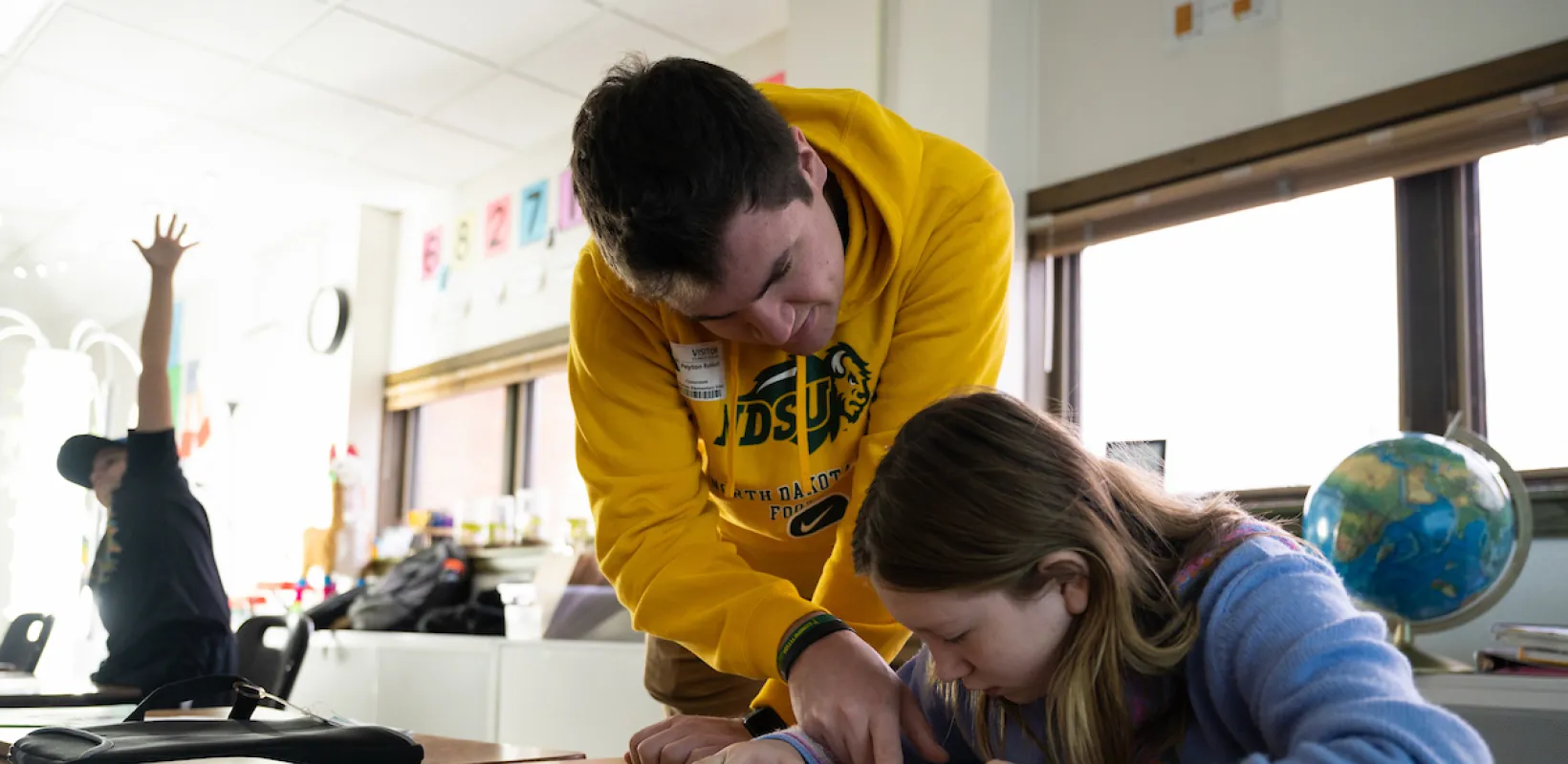 Student teacher helping a student at her desk with student in background raising arm enthusiastically.
