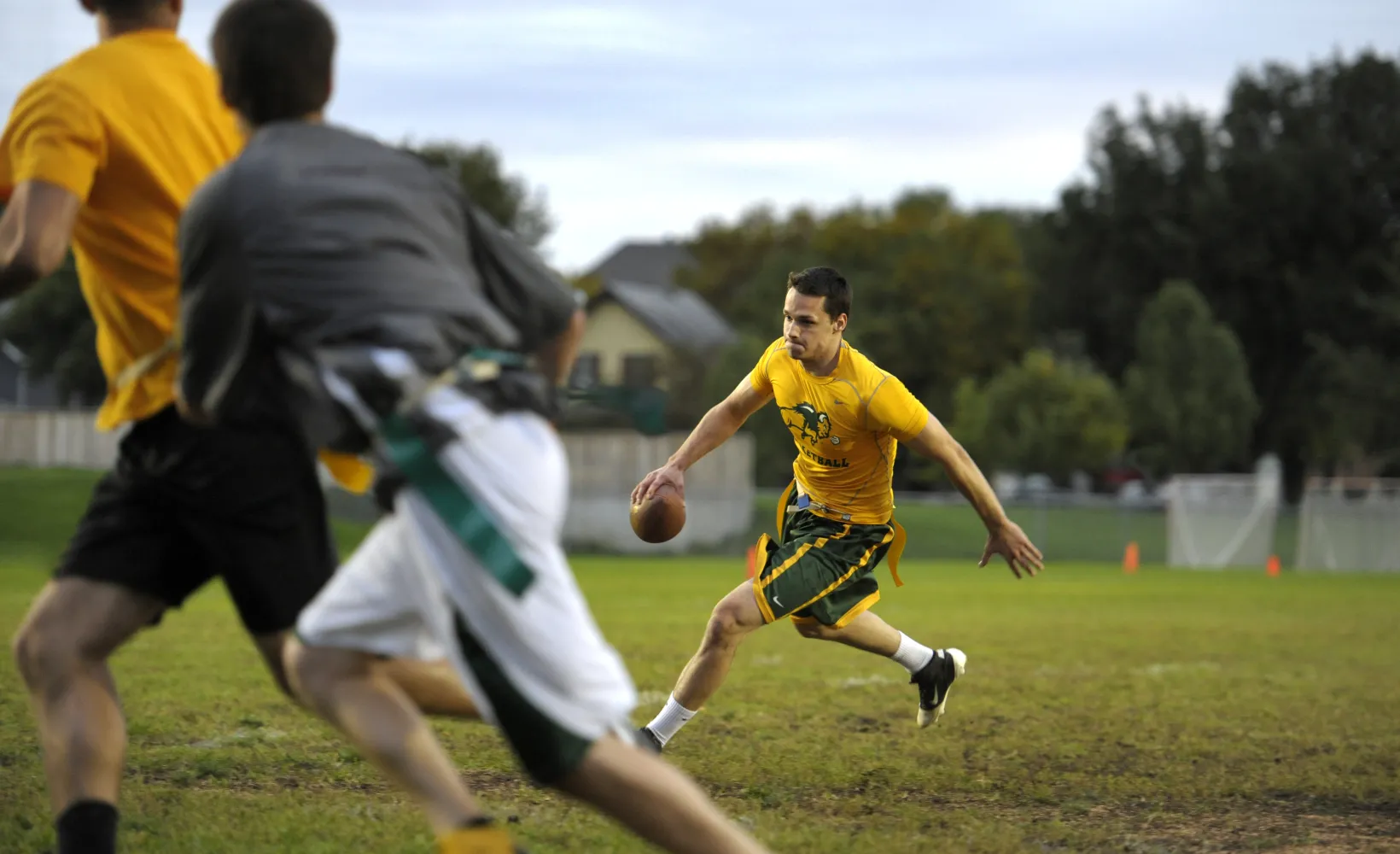 male students in green and yellow athletic gear running on a field holding a football