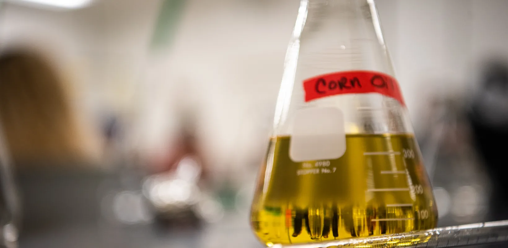 Glass flask containing yellow liquid identified as 'Corn Oil' sitting on a table.