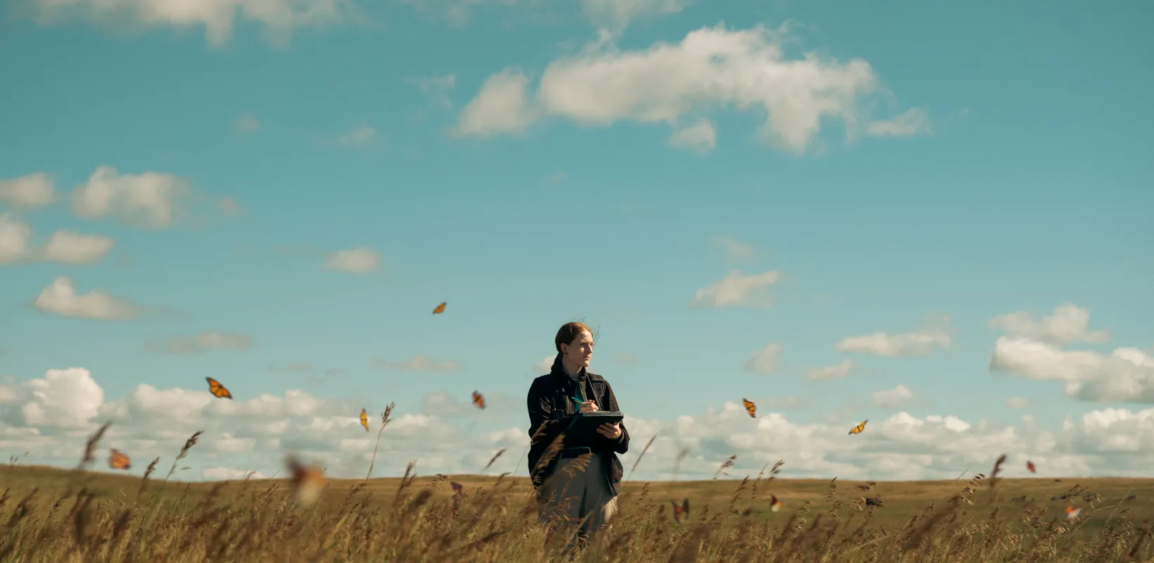 Student standing in a grassy field on a sunny day with Monarch butterflies flying above the grass.