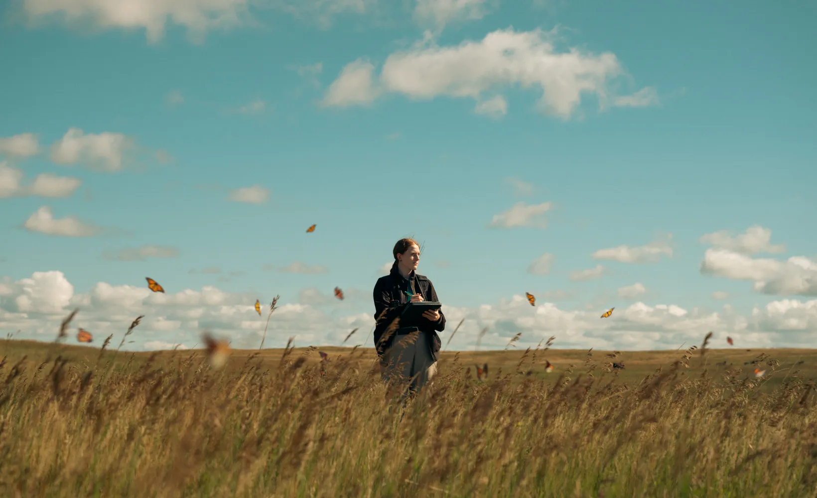 Student standing in a grassy field on a sunny day with Monarch butterflies flying above the grass.