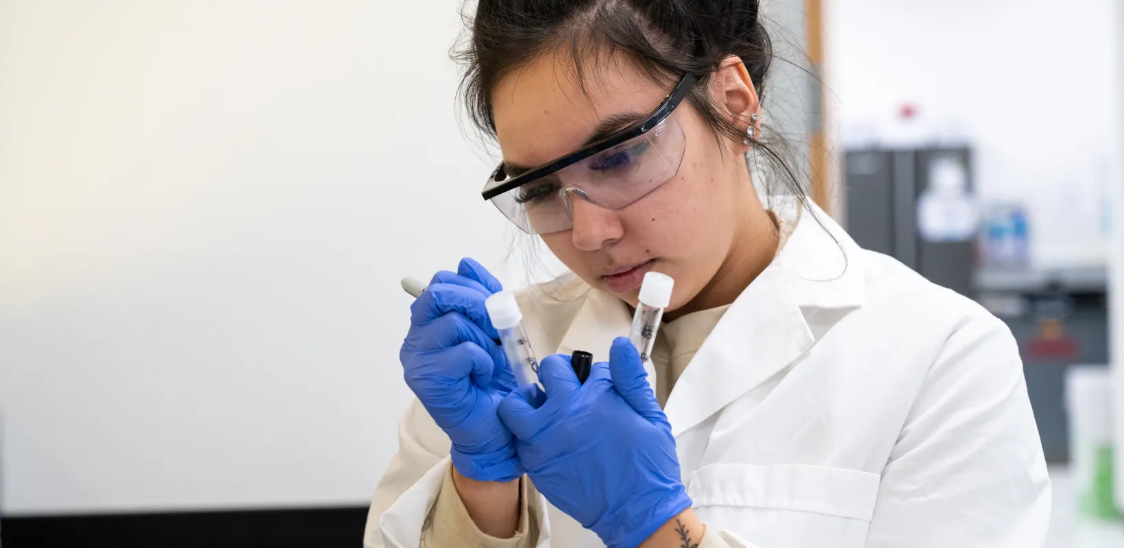 Student researcher working in the lab wearing safety glasses, lab coat and blue gloves while writing on a test tube with a sharpie marker.
