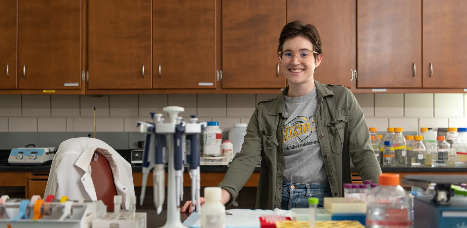 Student smiling confidently into the camera. She is standing in a lab wearing an NDSU shirt and green jacket.