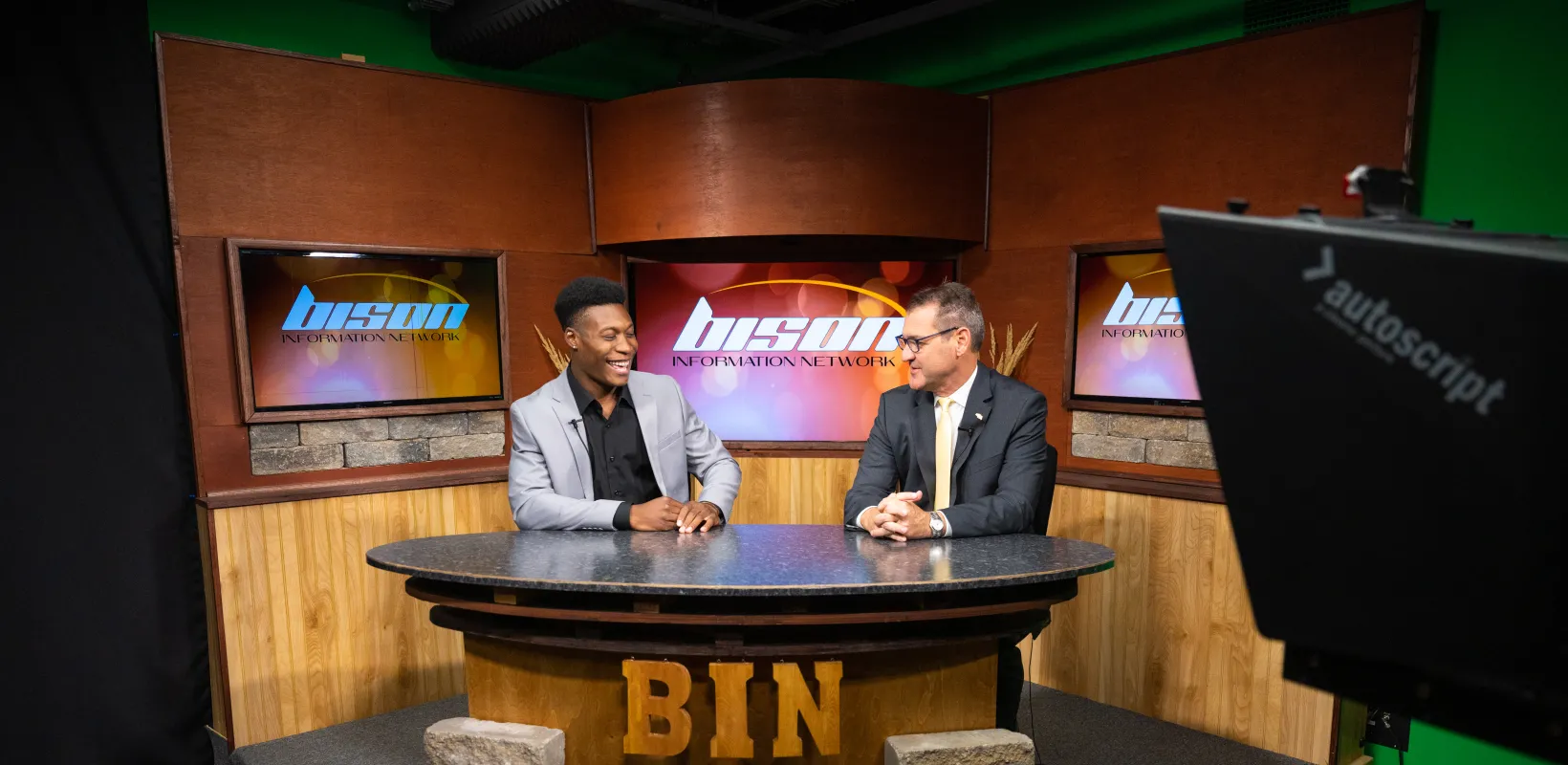 Student and President Cook sitting in the Bison Network studio having a conversation. Both wearing suits and smiling at one another. 