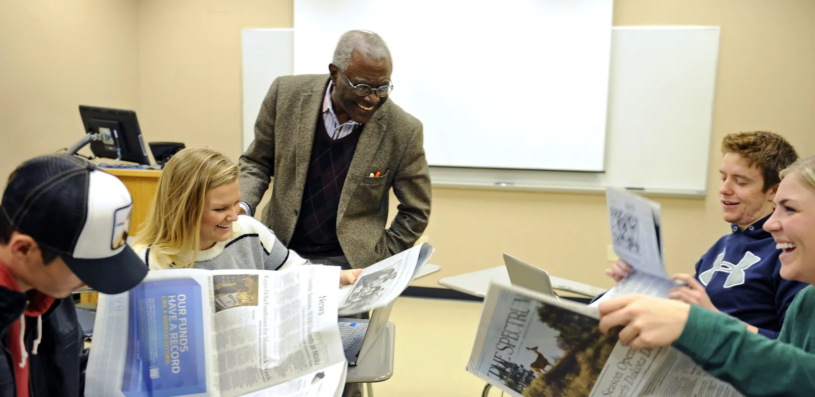 Students working with newspapers in a classroom setting. Professor and students laughing while looking at newspapers.