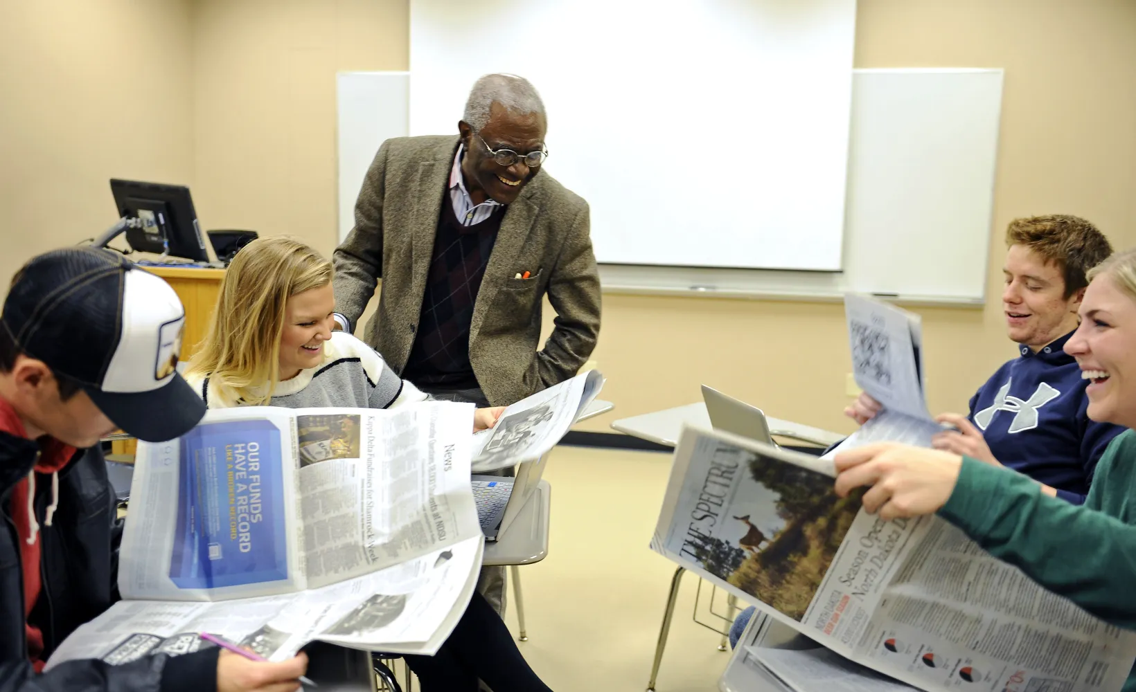 Students working with newspapers in a classroom setting. Professor and students laughing while looking at newspapers.