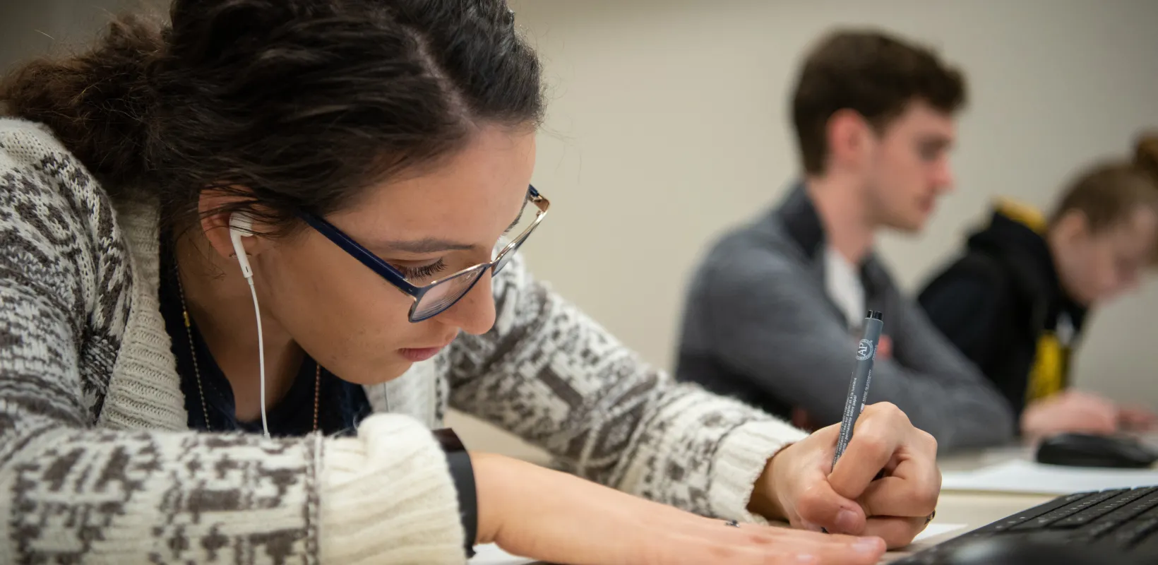 Student working at desk with pen and paper. Student is wearing glasses and earphones.