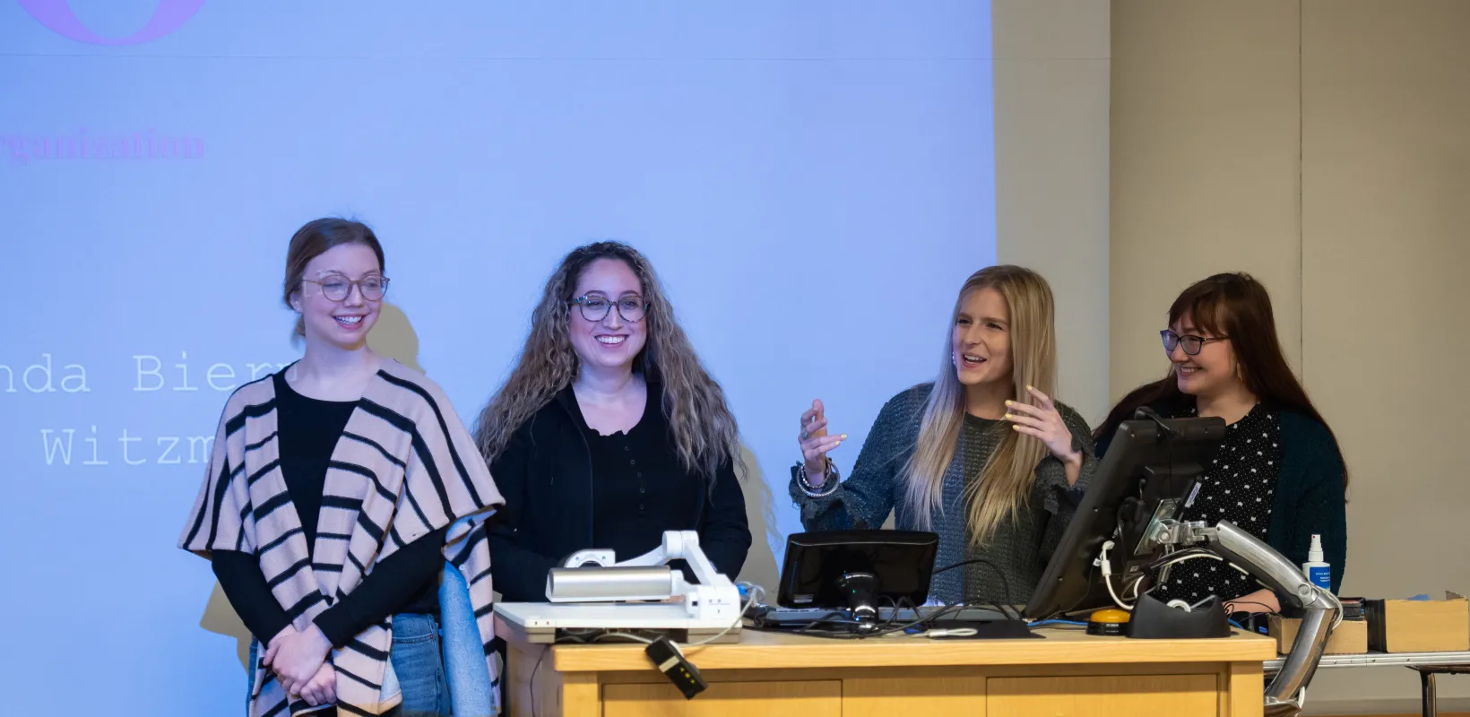 Four students presenting in front of a room. One student is speaking while the others smile confidently.