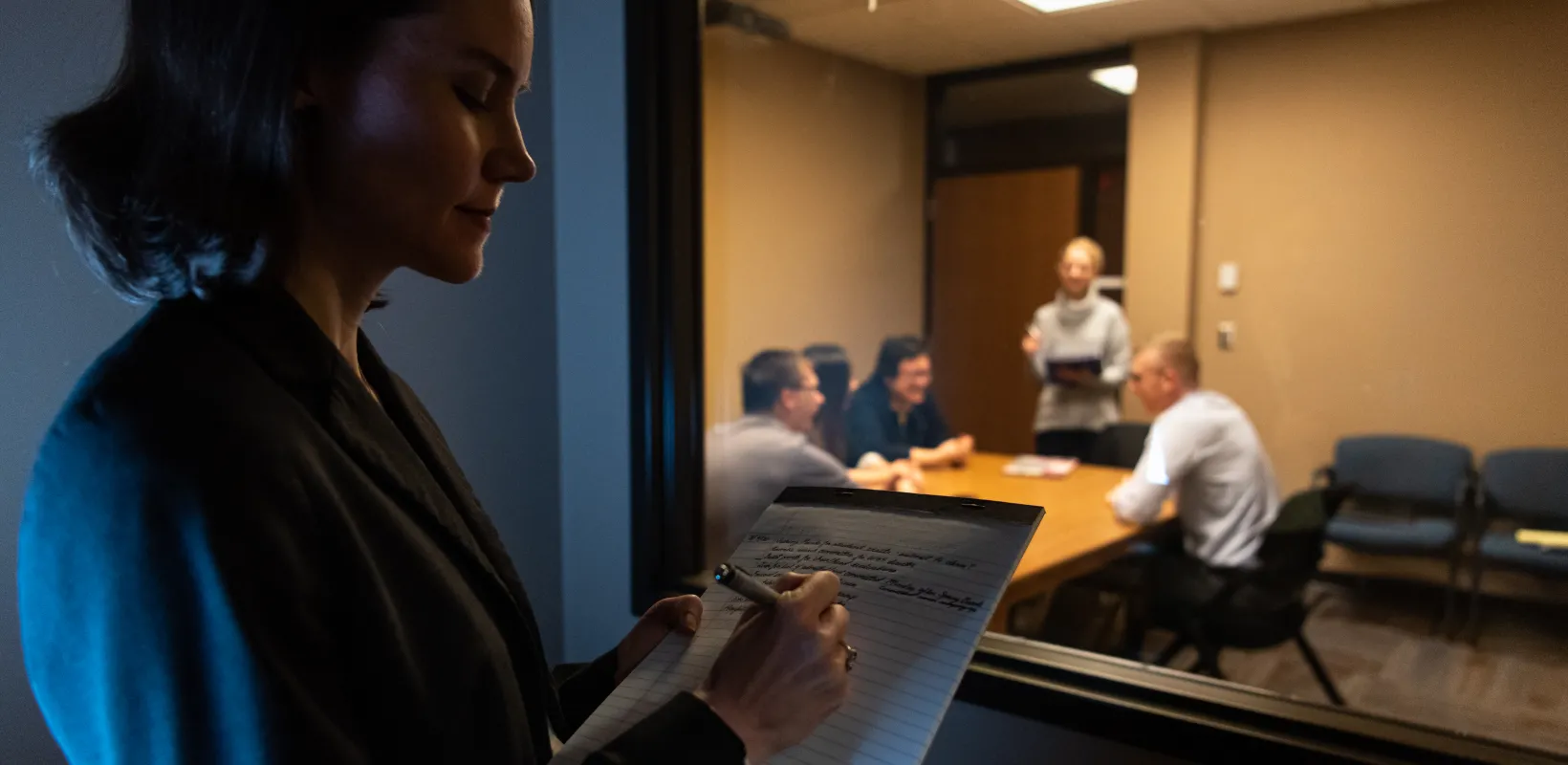 Faculty taking notes while observing a meeting through a window.