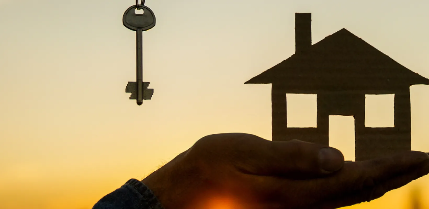 Person holding a model house with a key hanging above their wrist against a sunset backdrop