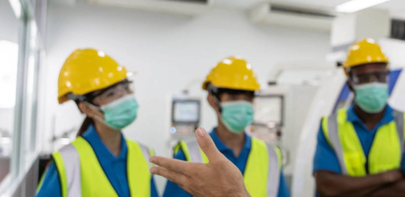 Three employees getting safety instructions in a factory, all have hard hats, safety goggles, reflective vests and masks on. 