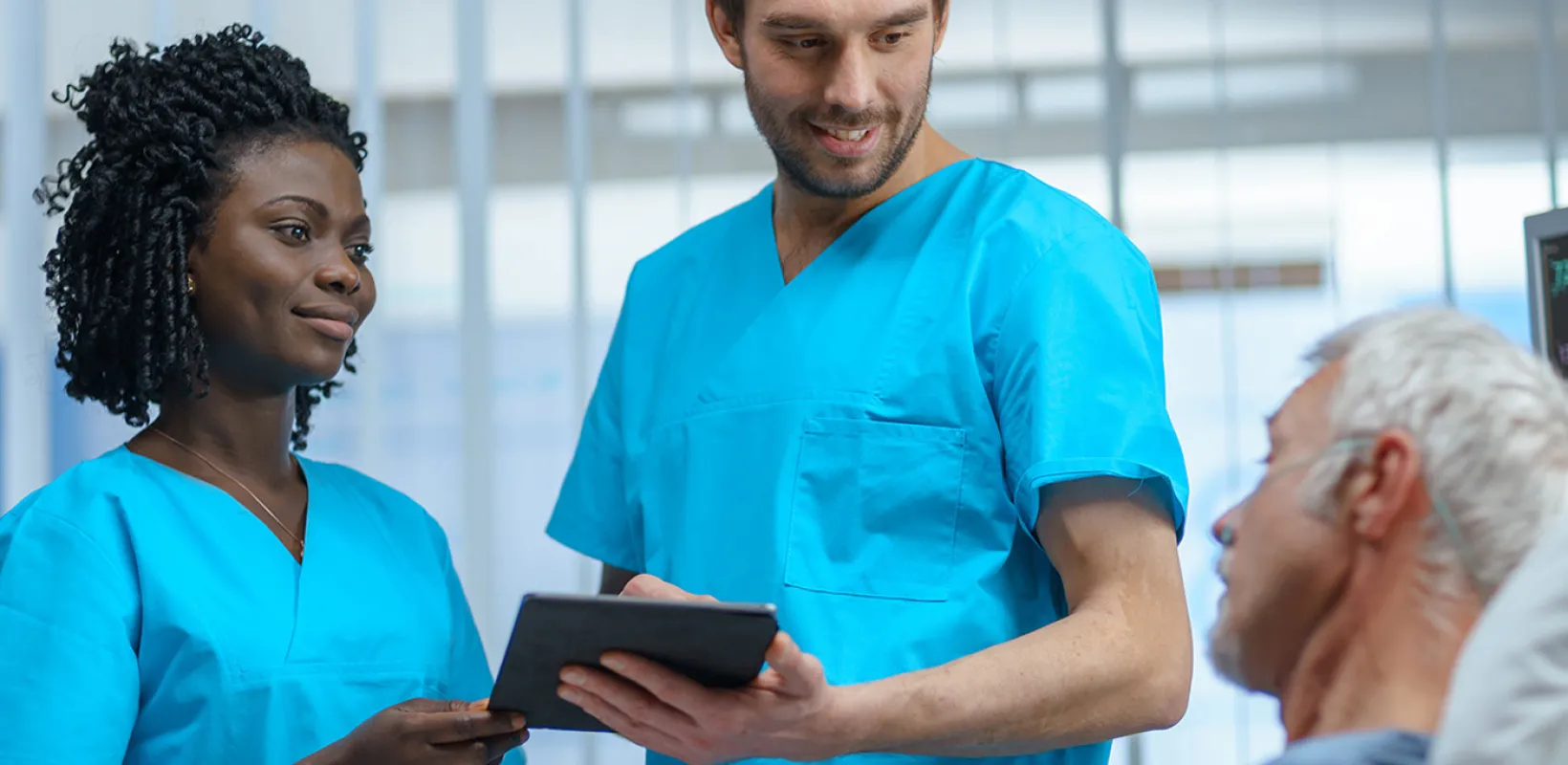 Two patient care technicians in nursing uniforms discussing a care plan while holding a tablet device