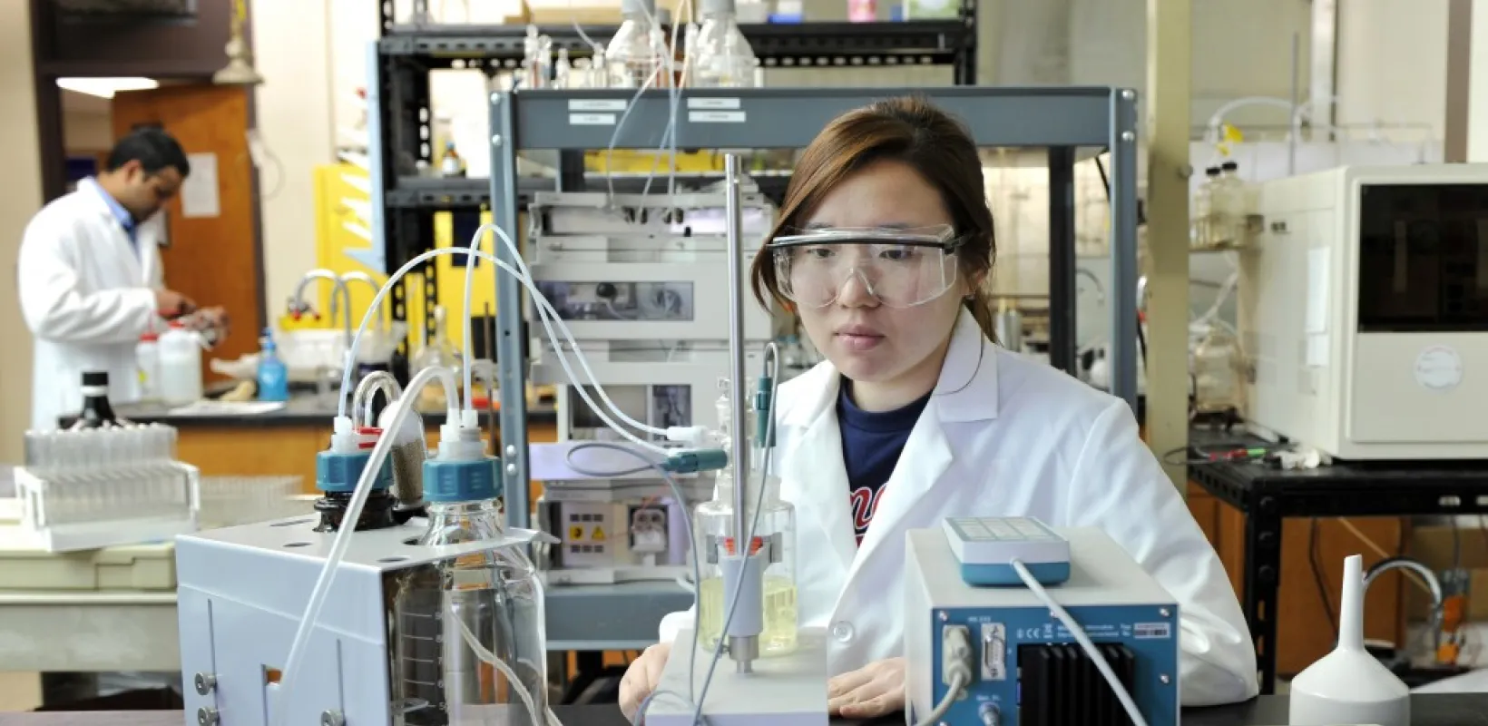 Research scientist wearing a white coat and protective eyewear while working in lab with lab equipment.