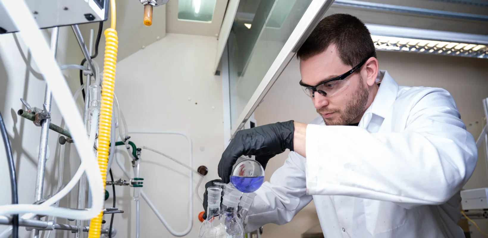Student researcher working in the lab wearing safety glasses, lab coat and black gloves while pouring a blue liquid from a beaker into a bottle.
