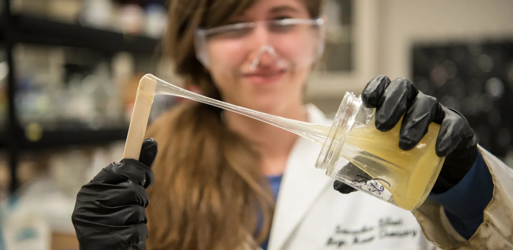 Research student wearing a white lab coat and black gloves while working with a yellowish, rubber substance in the lab.