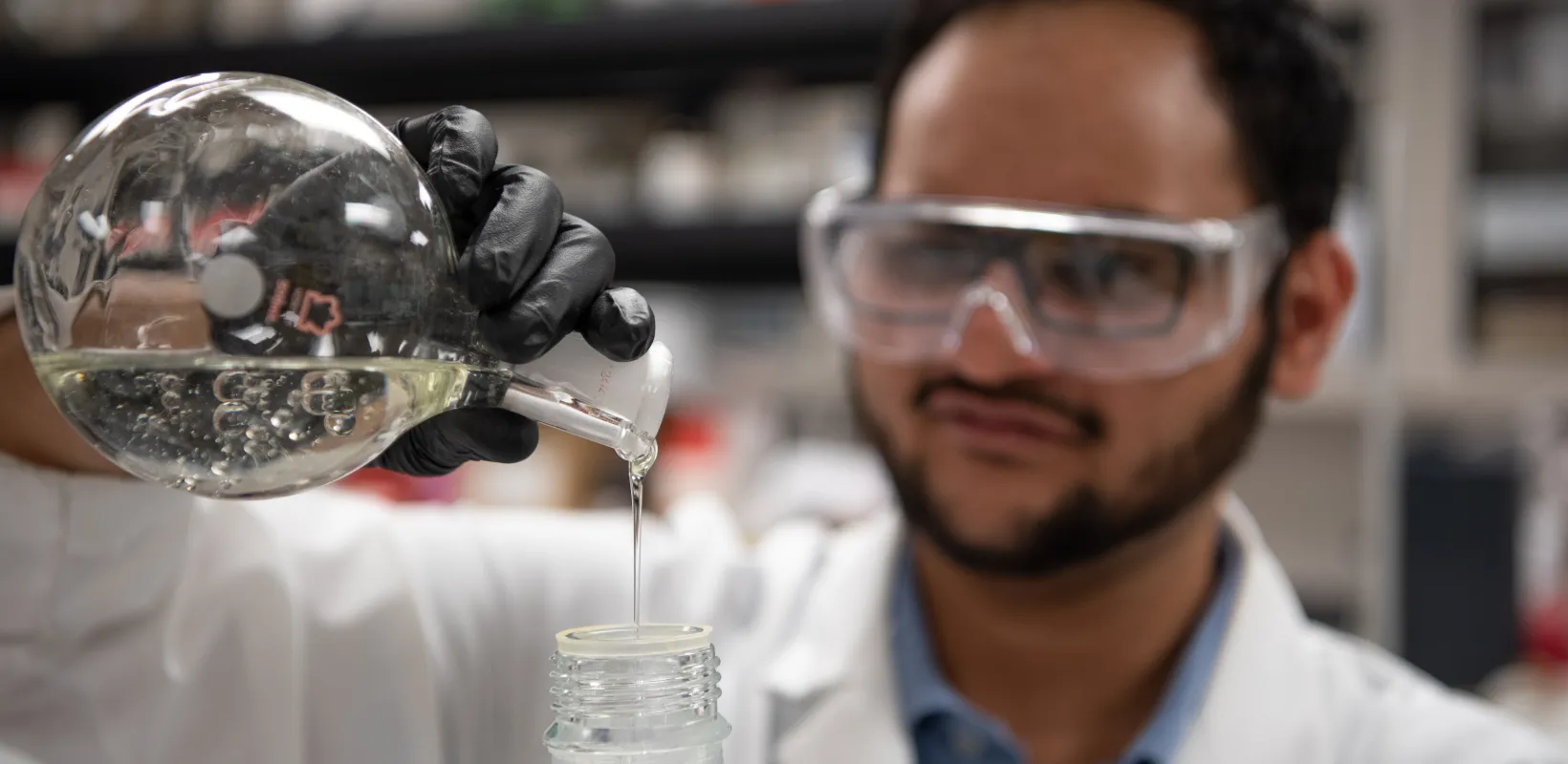 Student researcher working in the lab wearing safety glasses, lab coat and black gloves while pouring a clear liquid from a flask into a bottle.