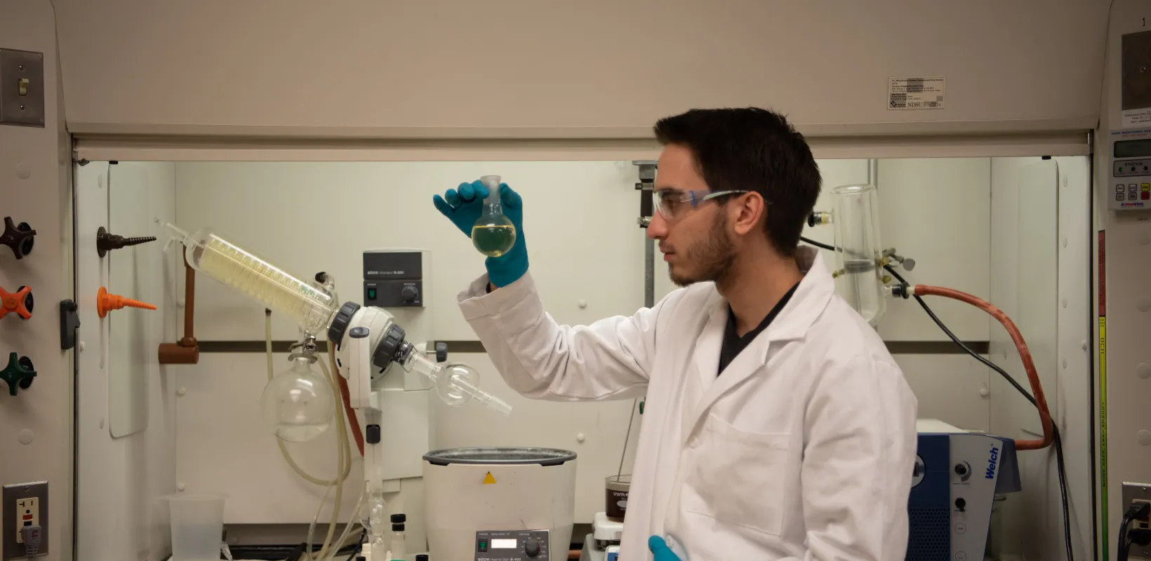 Student wearing a white lab coat and blue gloves looking at a clear yellow liquid in a glass flask.