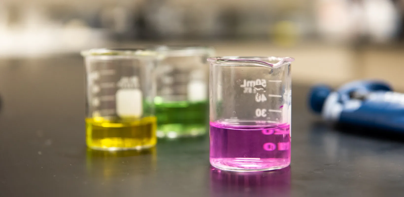 Three glass beakers sitting on tabletop containing a pink, yellow or green liquid. 