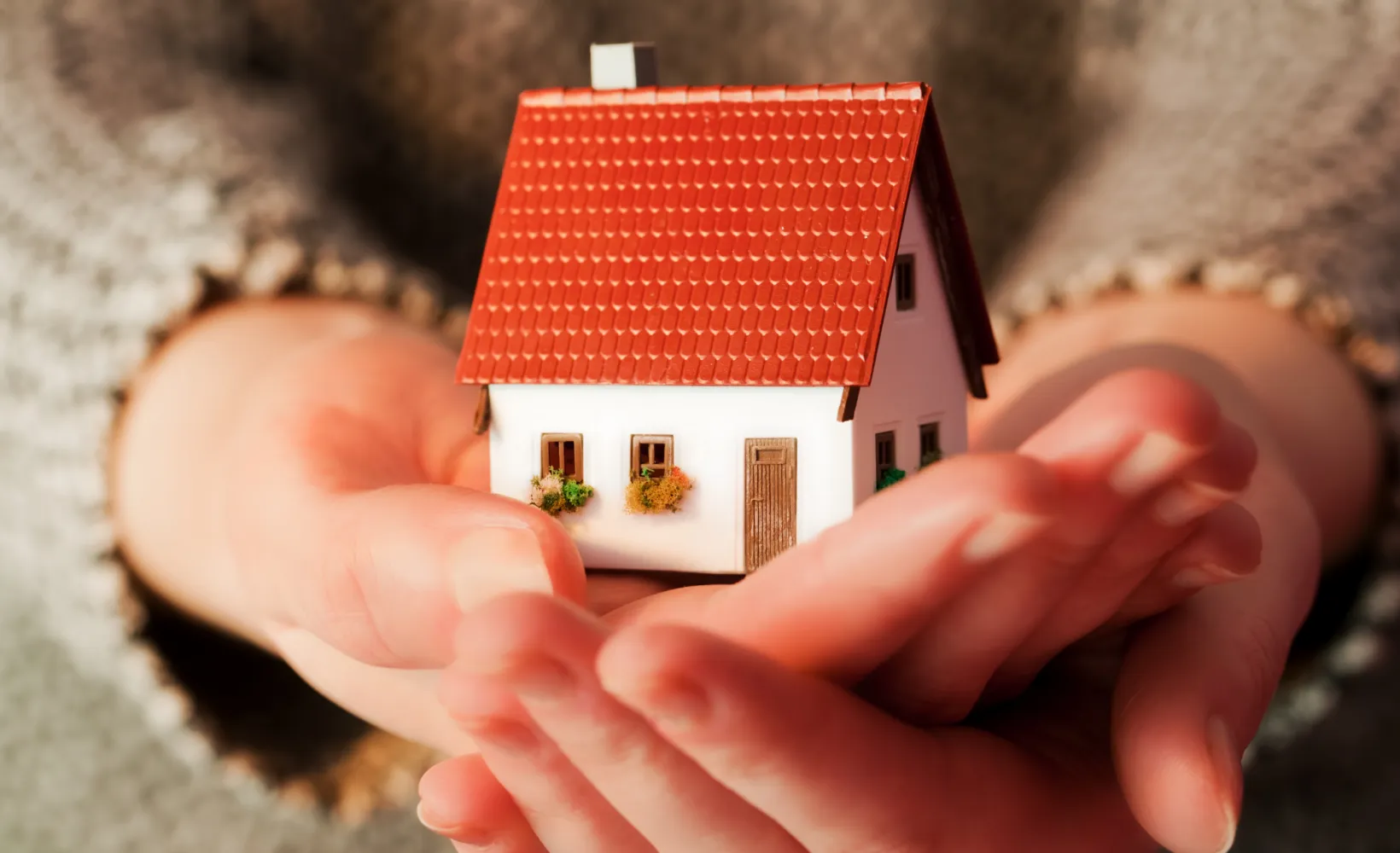 Hands holding a small plastic, white house with a red roof.