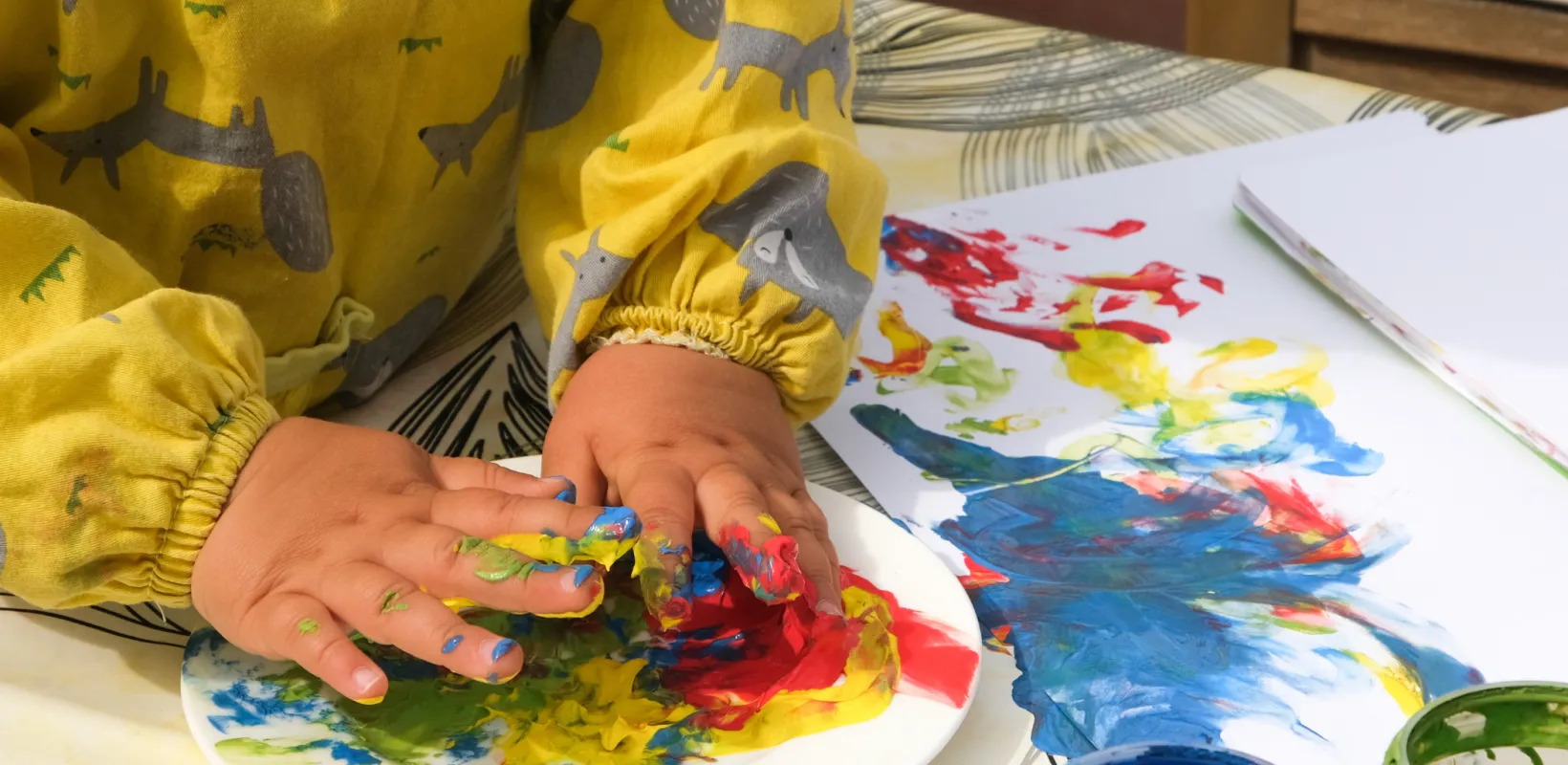 A child wearing a yellow long-sleeved shirt working with finger paint and white paper.