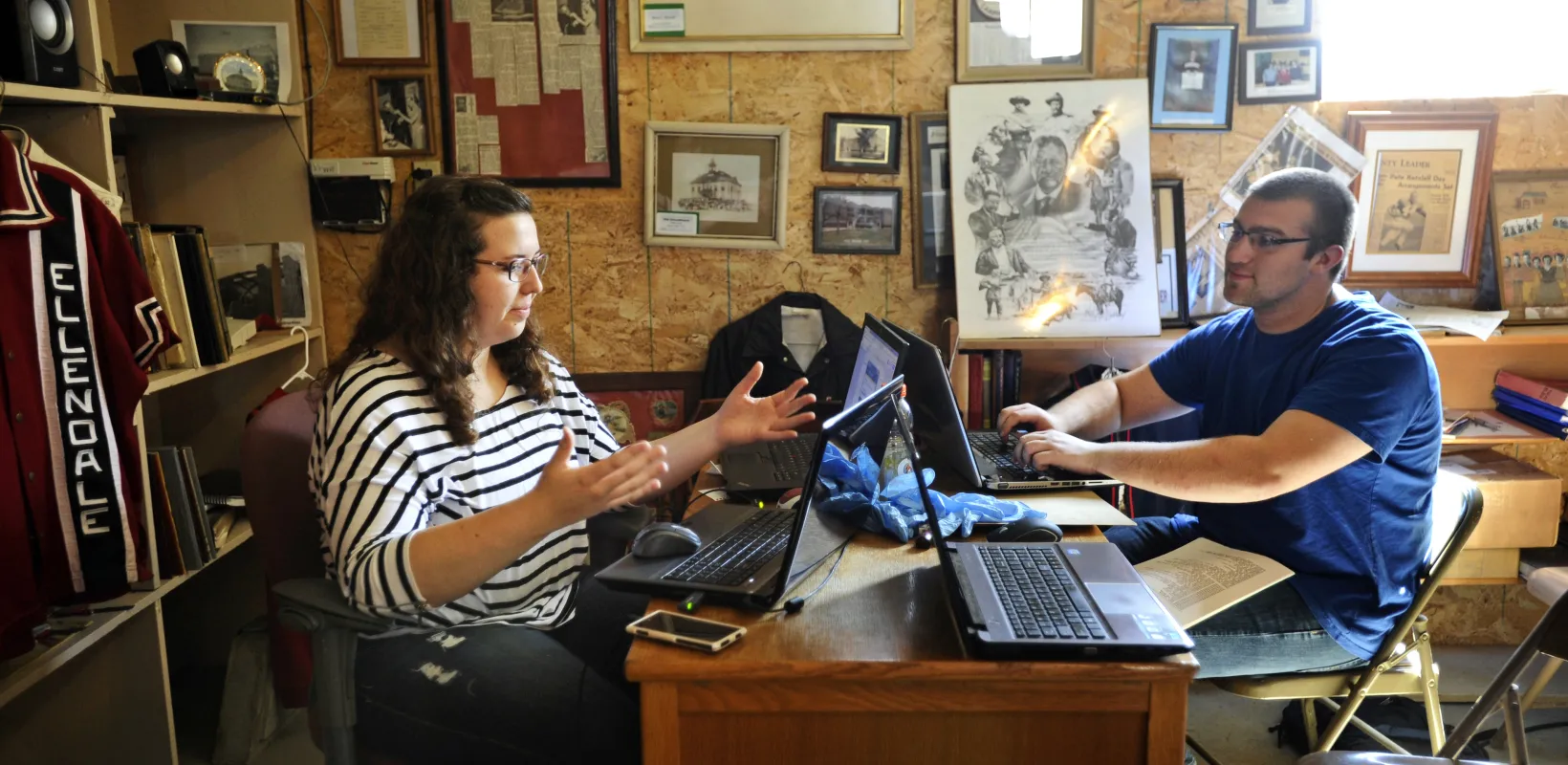Students sitting at desk with laptops while one student is talking and the other is listening.