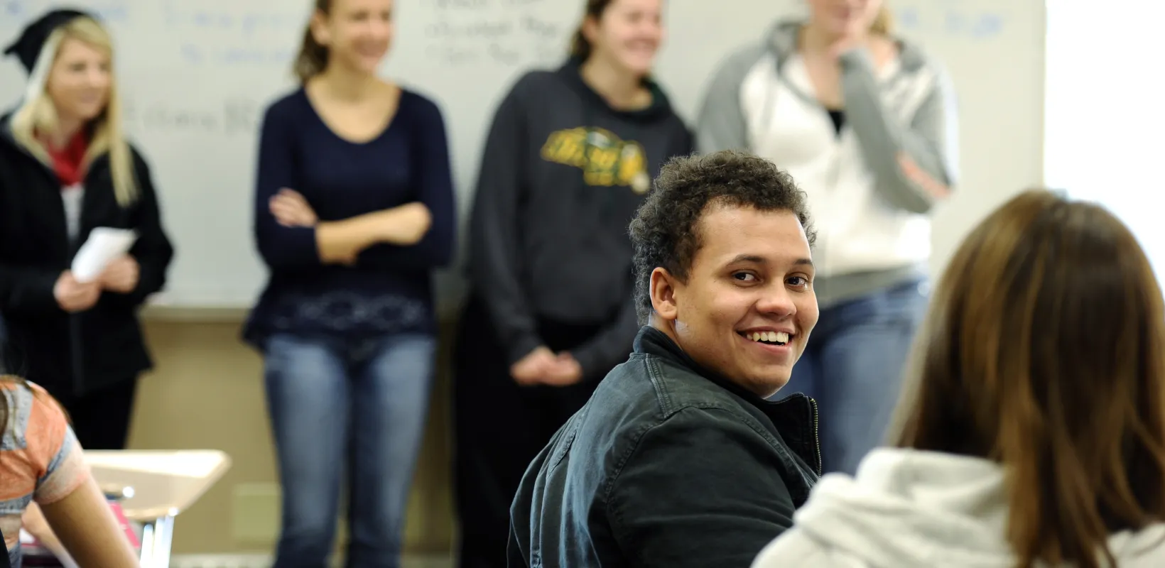 Student smiling at camera in a classroom setting with other students in the background.