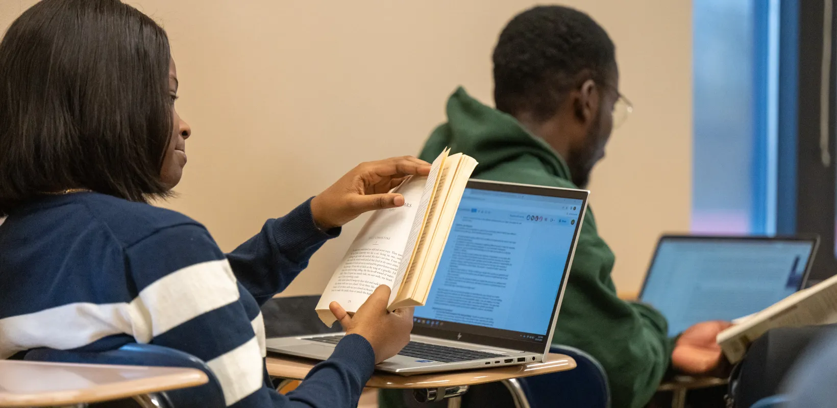 Studing reading a book while sitting at her desk with her laptop. 
