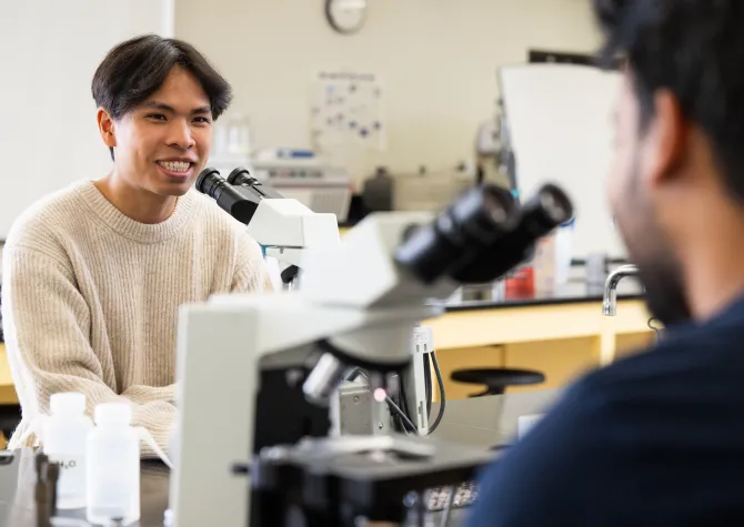 A student chats in the lab with another student also sitting at a microscope in the foreground