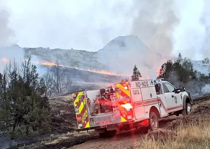 White fire engine with flashing red lights parked near a hill that is on fire with smoke trailing up into the sky