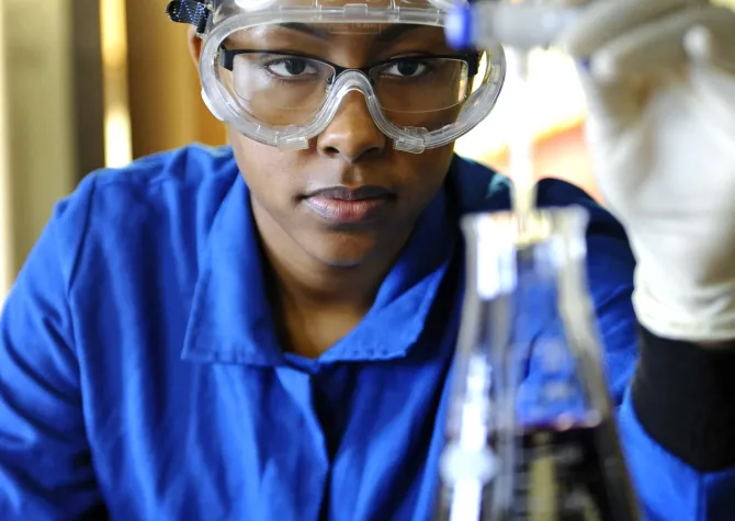 Student researcher wearing a blue shirt and lab googles looking intently at a beaker containing a dark purple liquid