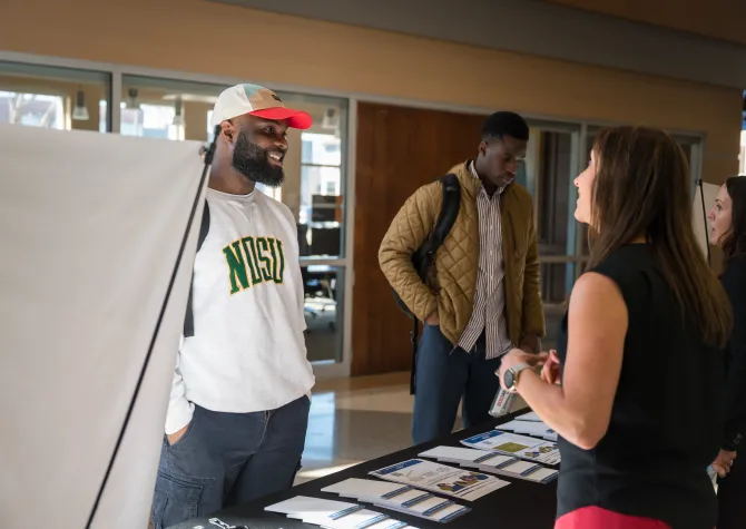 Male student speaking to a woman at a booth in an academic building 