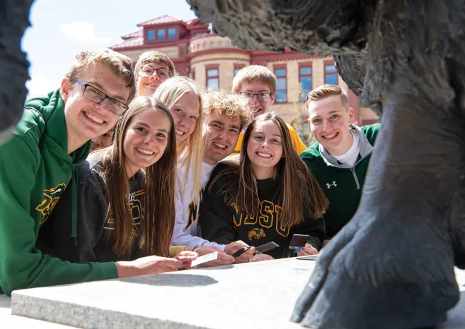 Eight students pose behind front hoof of Bison statue.