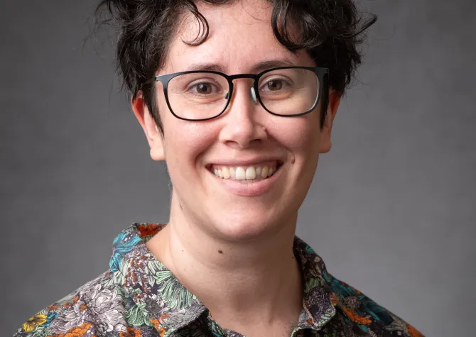 Evelyn Milburn, a white woman with brown hair and glasses is wearing a bright collared shirt and smiling broadly.