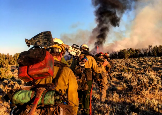 A line of Wildland firefighters walk forward as black smoke rises in the distance