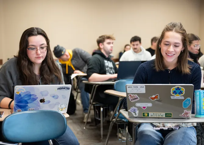 Students working on laptops while sitting in desks located in classroom.