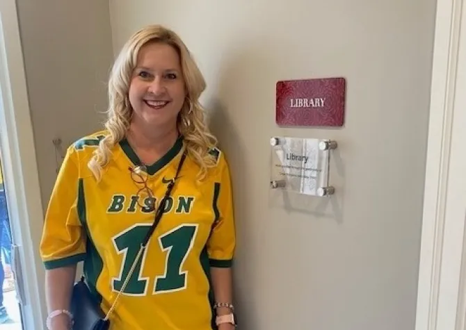 a woman wearing an NDSU jersey stands by a sign that says "library"