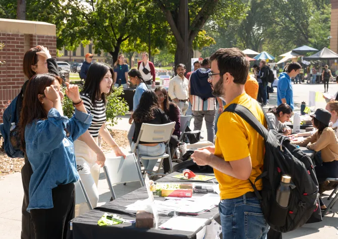 Students gather at an outdoor campus involvement fair or club recruitment event where tables are set up for student organizations