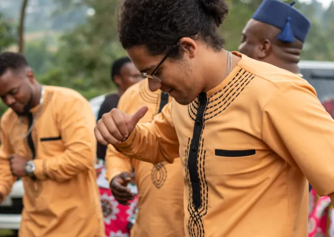 Kingston Kamangala, a Black man with curly hair and glasses, smiles as he dances in a yellow tunic.