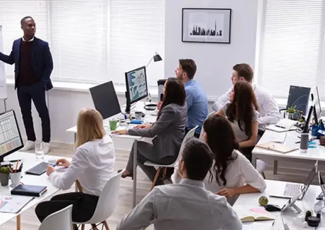 Group of workers at desks with computers listening to leader pointing at poster with metrics on it. 