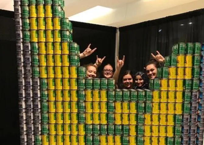 ASID students posing in front of a image created out of black, green and yellow cans.