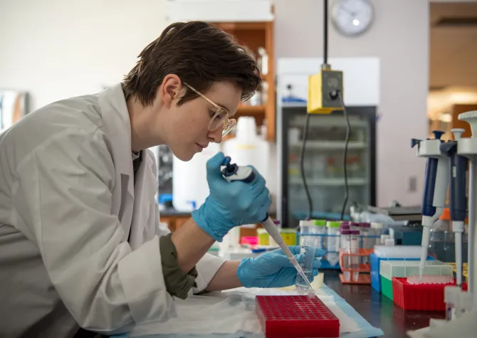 Elena working in a lab setting wearing a lab coat and gloves while holding a pipet.