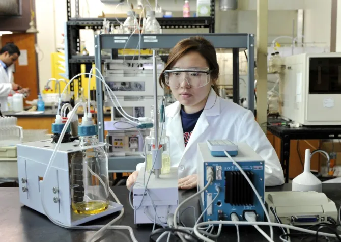 Research scientist wearing a white coat and protective eyewear while working in lab with lab equipment.