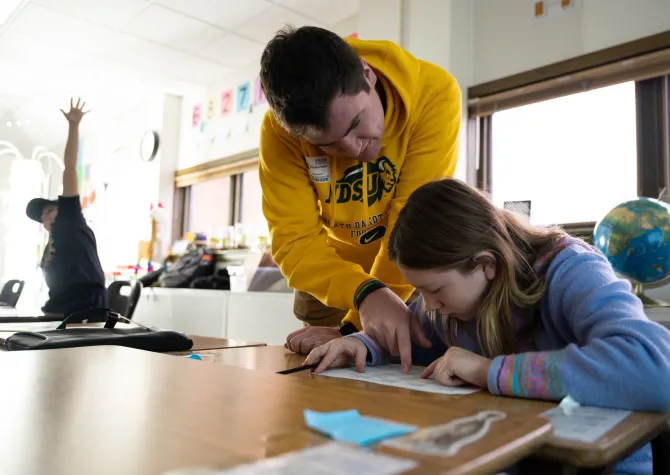 Student teacher leaning on student desk explaining and pointing at students paper.