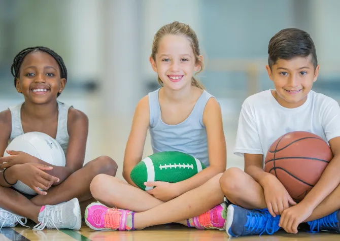 Three young students sitting cross-legged, holding different types of ball, smiling at the camera.