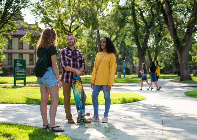 3 NDSU students standing on sidewalk visiting. Background inlcudes green leafy trees, green grass and students walking in the distance.