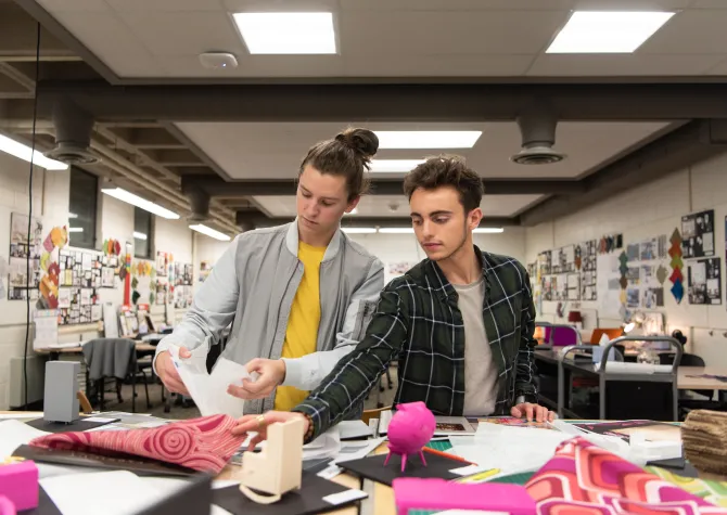 Two students working at a table covered in fabric, paper, and other interior design related tools. 